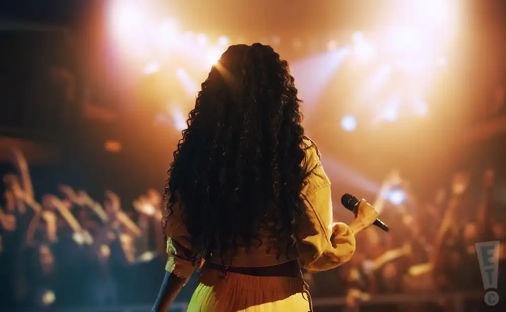A singer with long, dark curly hair stands with her back to the camera, holding a microphone on a brightly lit stage.