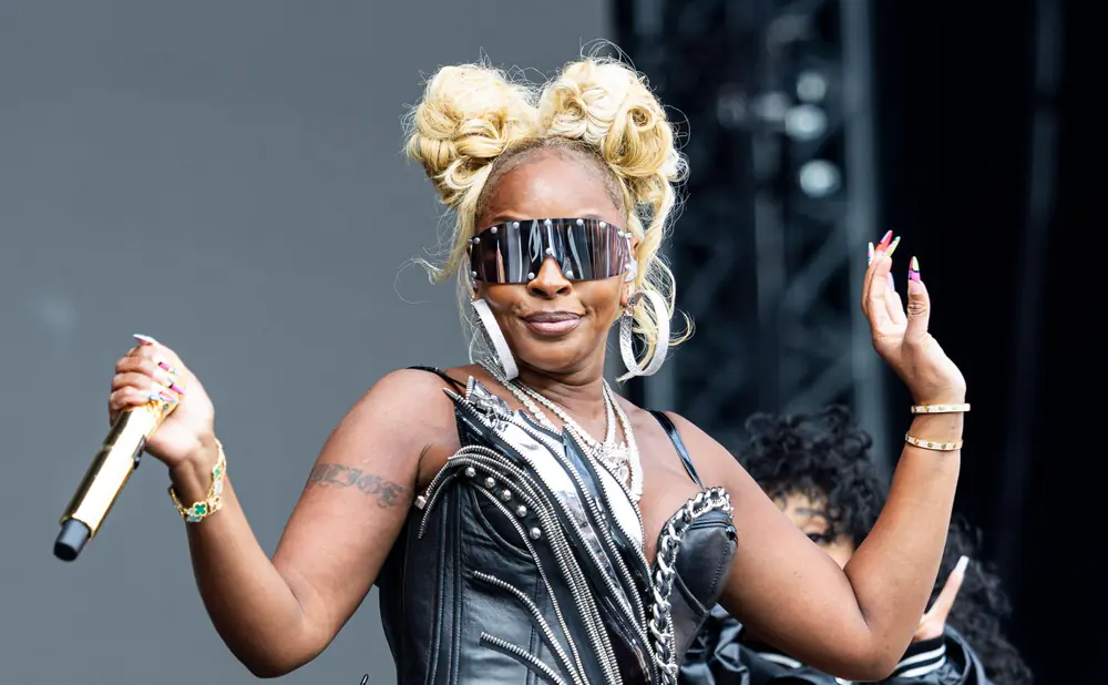 Mary J. Blige in a black leather outfit, sunglasses, and blonde hair in buns, holding a gold microphone.