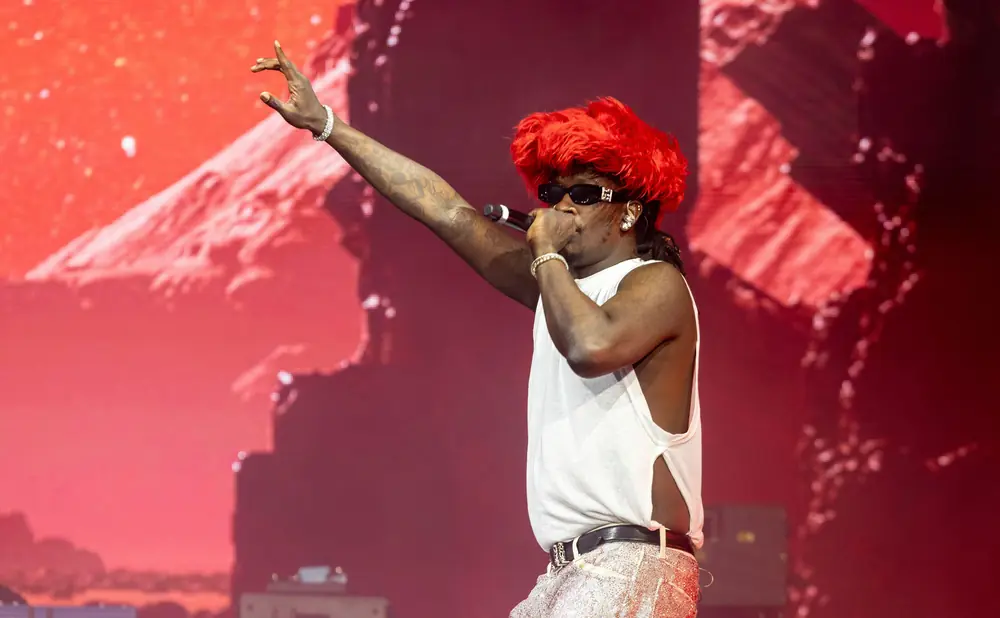 Rapper with bright red hair and sunglasses performs on stage in a white tank top and sparkly pants.