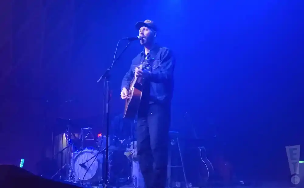 A man in a blue denim jacket and baseball cap sings and plays an acoustic guitar on a stage lit with blue light.