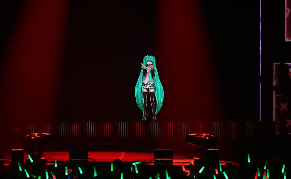 A digital anime character with long teal hair performs on a stage lit by red and green lights, with an audience holding glow sticks.