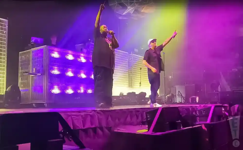 Two men perform on a stage lit with purple and yellow lights, with a DJ booth behind them.