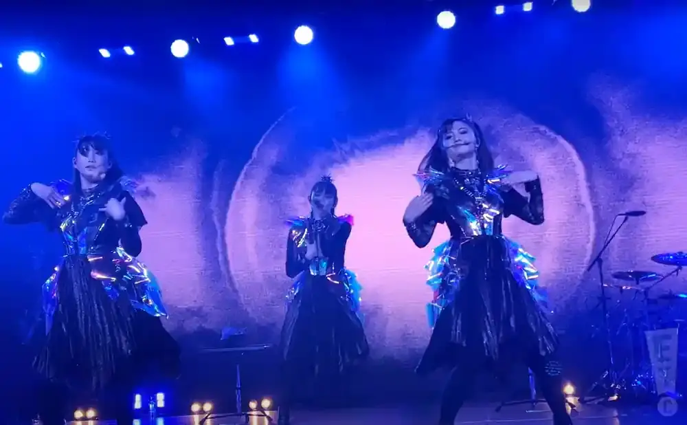 Three women in sparkly, futuristic outfits perform on a stage with blue lighting.