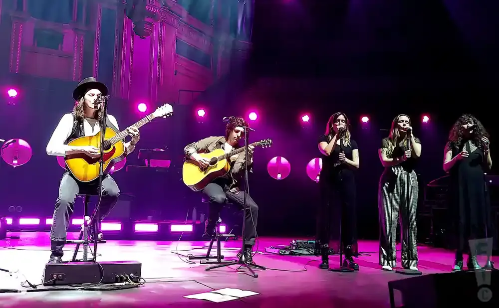 Musicians perform on a stage with purple lighting. A man in a hat plays an acoustic guitar.