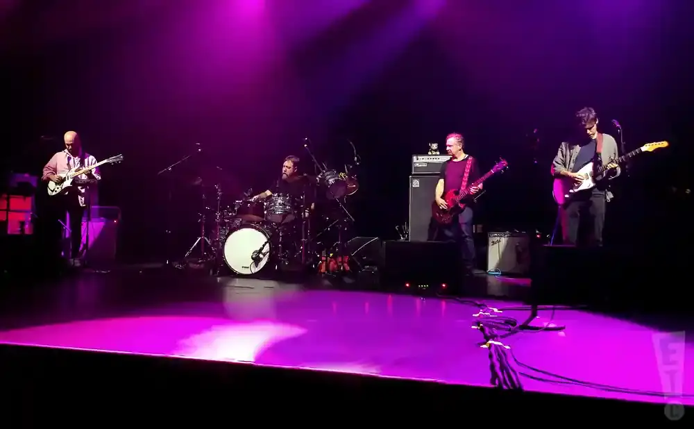 A band performs on a stage lit with purple lights. Four musicians are visible playing guitars and drums.