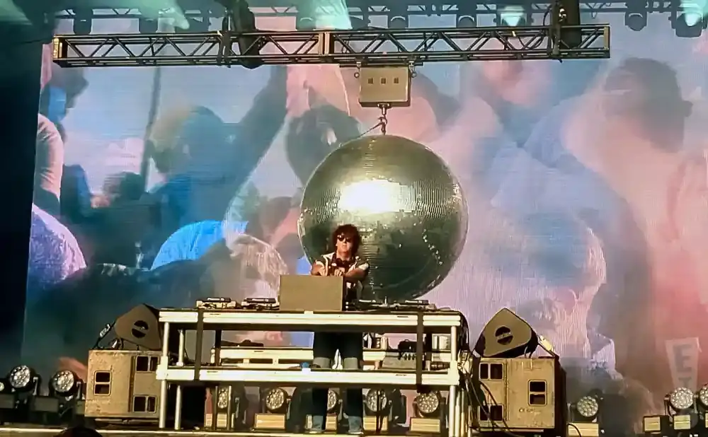 A DJ plays at a turntable under a disco ball with a blurred crowd in the background.