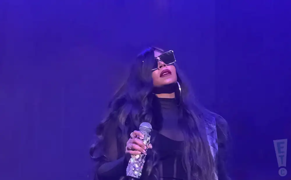 Singer with long dark hair, large sunglasses, and a bedazzled microphone performs on a purple stage.