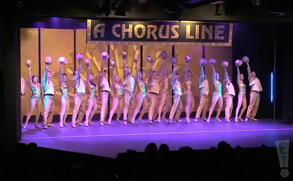 Dancers in sequined outfits and hats perform on stage in front of a 