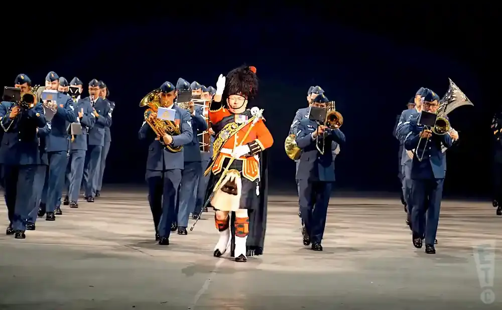 A marching band in blue uniforms and a leader in a red and black ceremonial uniform play instruments on a stage.