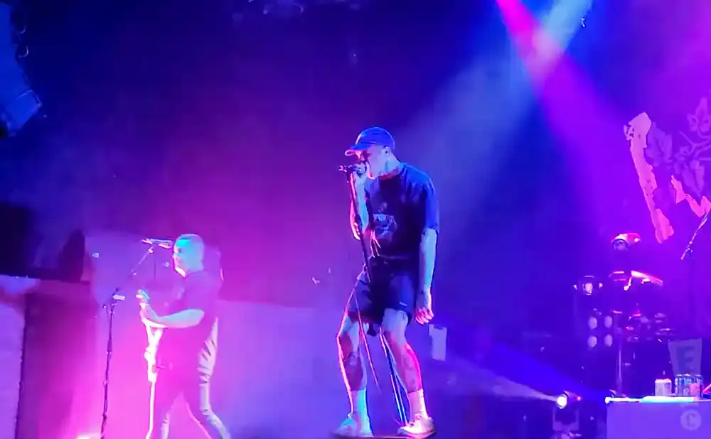 A male singer in a blue cap performs on stage with a guitarist to his left, under bright purple and blue lights.