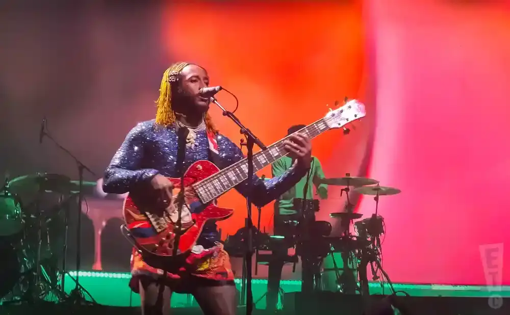 A guitarist with yellow dreadlocks plays a red guitar on a stage with a pink backdrop.
