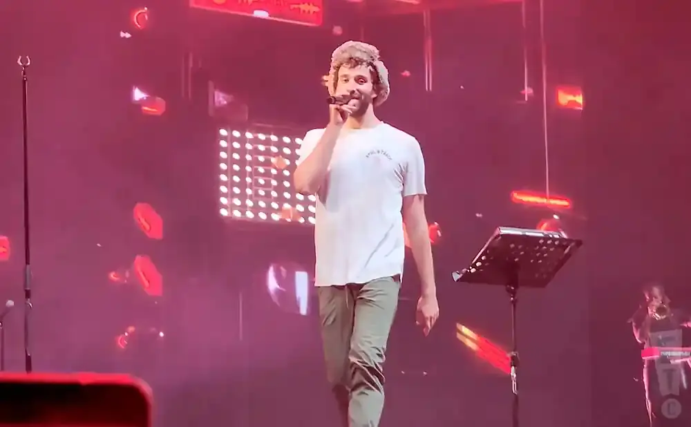 A man in a furry hat sings into a microphone on a stage with pink lights.
