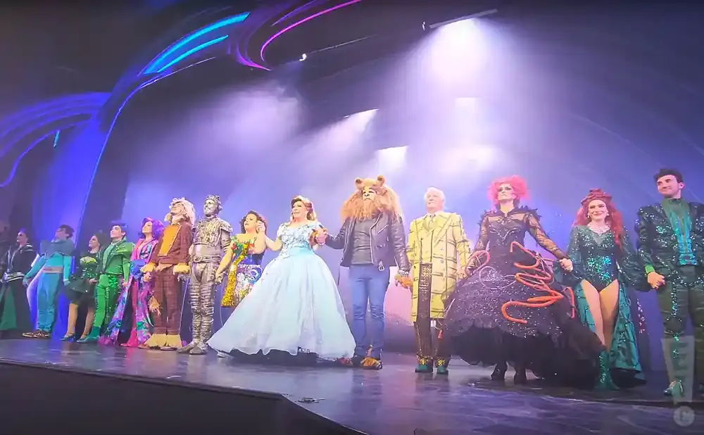 A cast in colorful costumes takes a bow on stage, with smoke and bright lights in the background.
