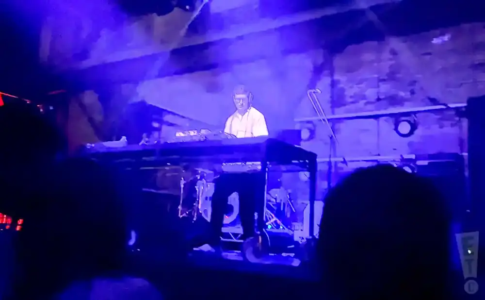 A DJ wearing headphones and a white shirt performs on a synthesizer in a dark, blue-lit venue.