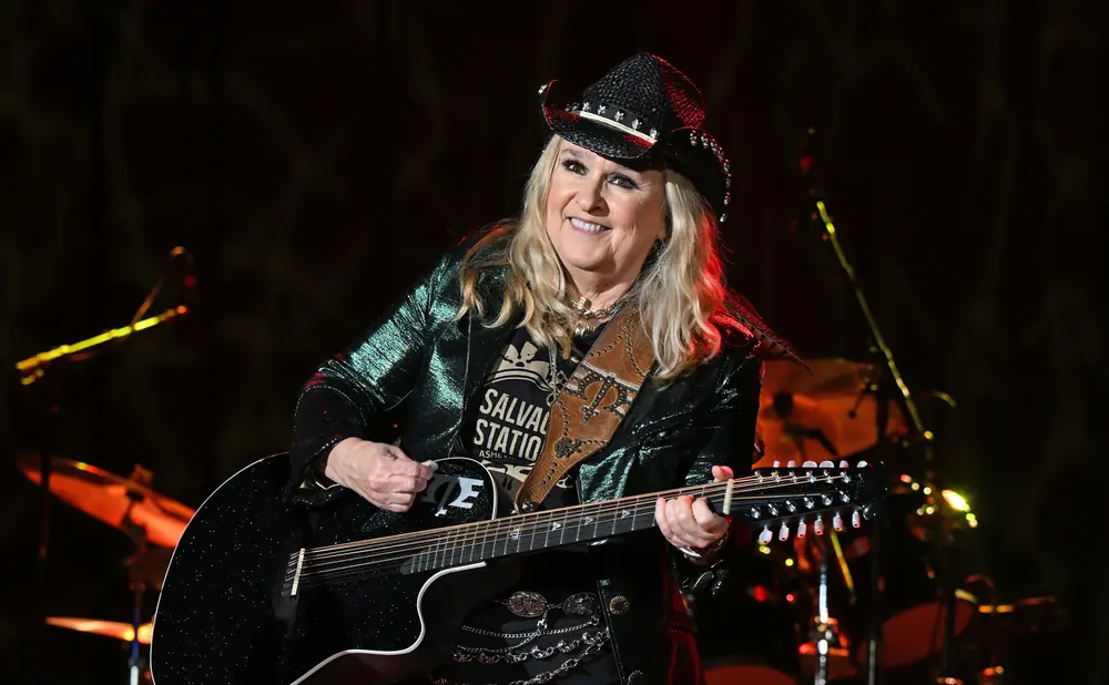 A blonde woman in a sparkly green jacket and cowboy hat plays a black 12-string guitar on stage.