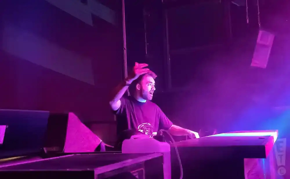 A male DJ with a beard, wearing a black t-shirt, performs on a stage with purple lighting.