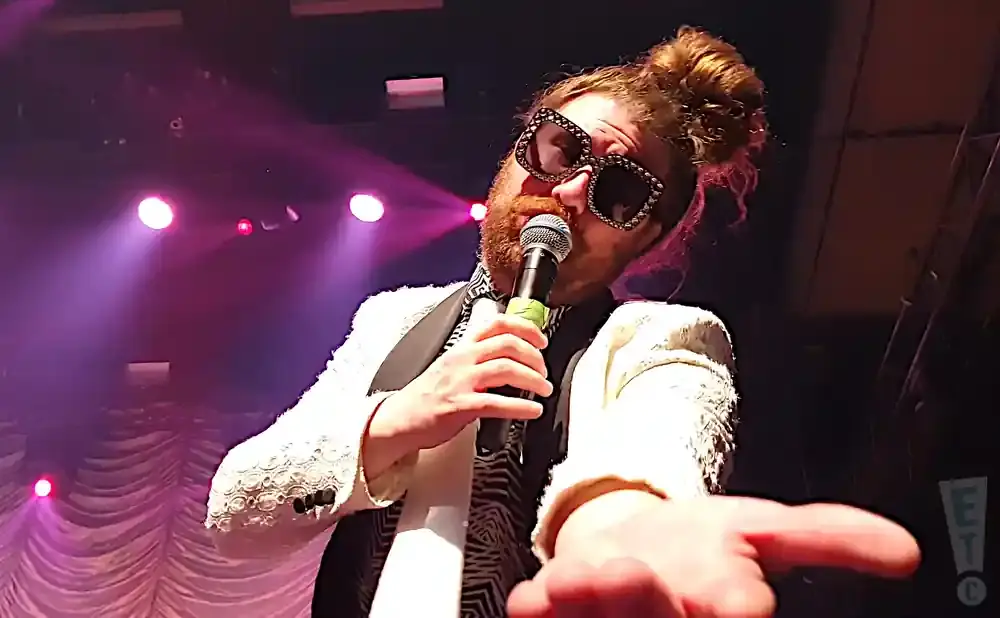 Man with messy bun, large bedazzled sunglasses, and a white jacket sings into a microphone on stage.