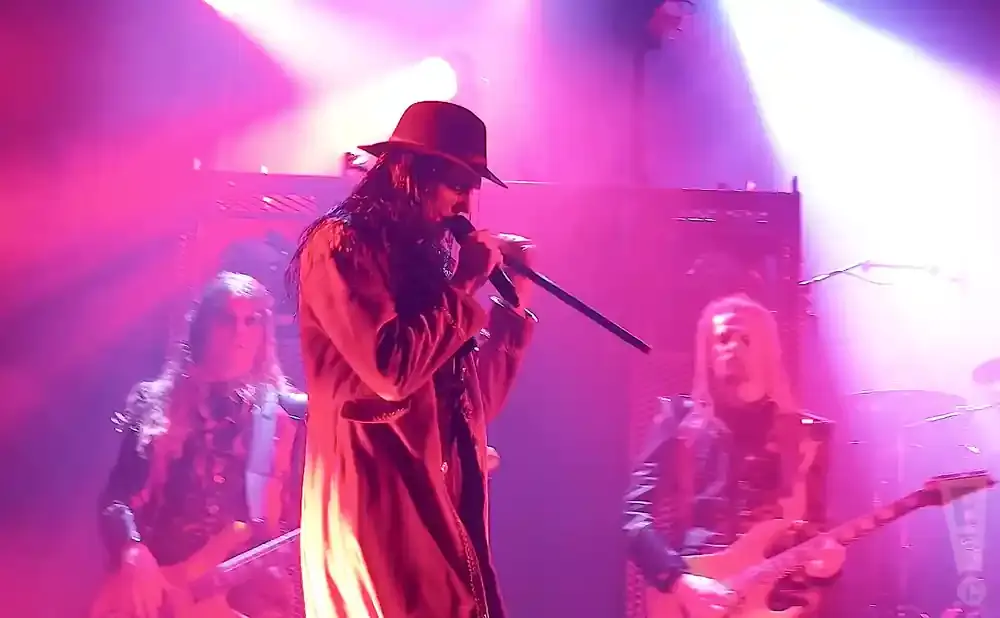 A band performs on stage with pink and purple lighting, a lead singer in a hat and long coat holds a microphone.