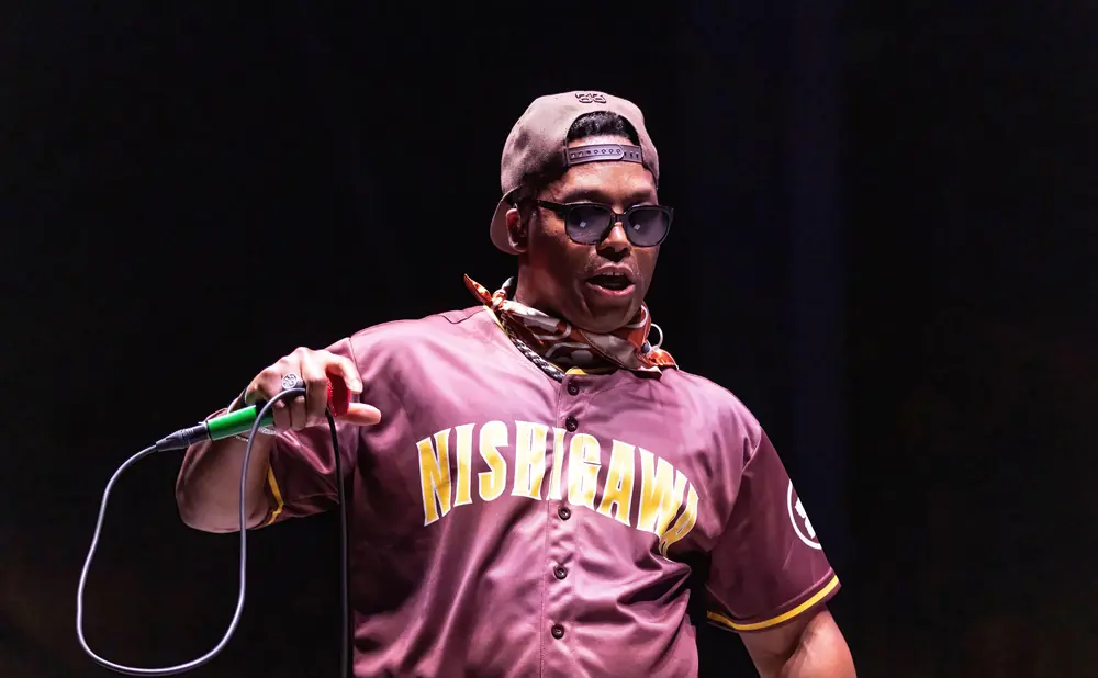 A Black man in a baseball jersey and sunglasses holds a microphone on stage.