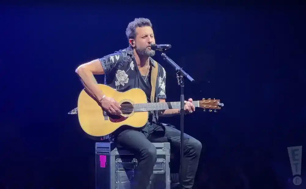 A man with a beard plays an acoustic guitar and sings into a microphone on stage.