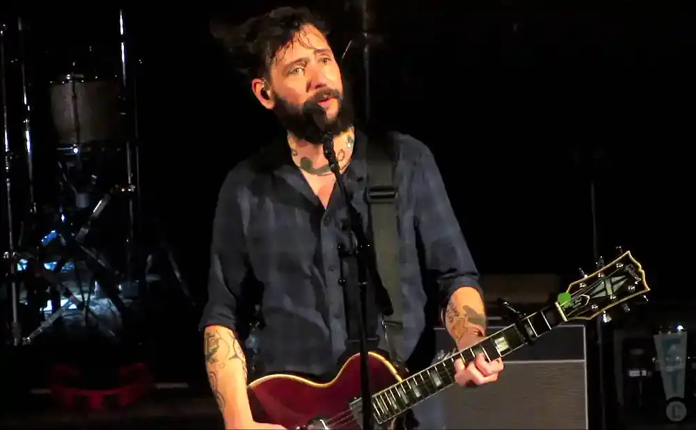 A man with a beard and tattoos plays a red guitar and sings into a microphone on a dark stage.