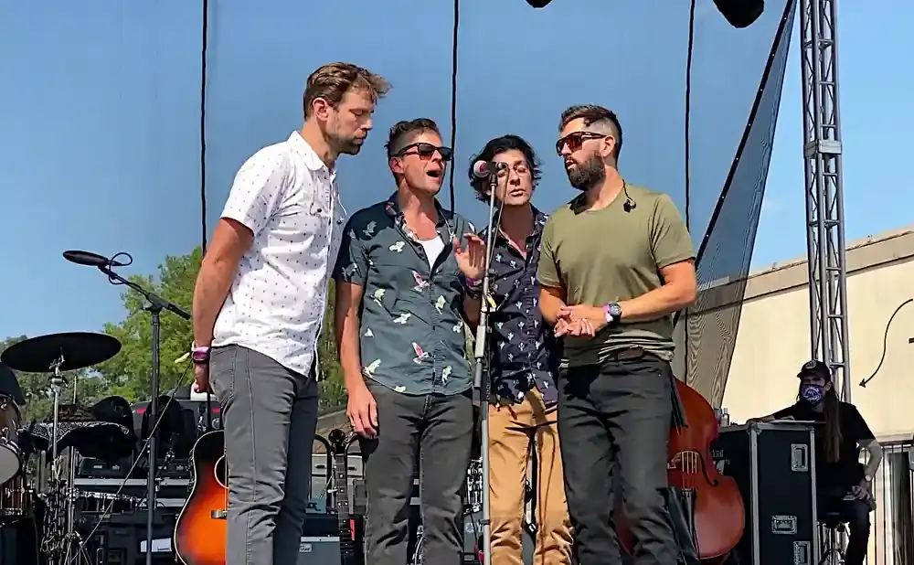 Four men in casual clothes sing into microphones on an outdoor stage.
