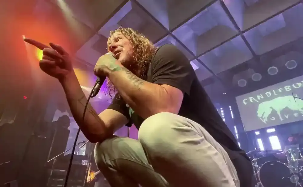 A male singer with long blonde hair crouches and sings into a microphone, pointing his finger towards the left.