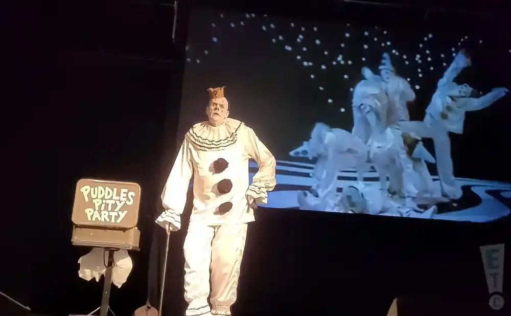 A clown with a question mark on his head stands on stage, holding a cane, with a sign that says 