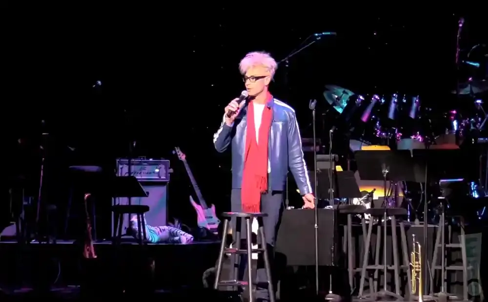 A man with blond hair and glasses, wearing a red scarf and blue jacket, holds a microphone on stage.