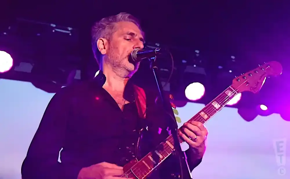 A man in a black shirt plays an electric guitar on a stage with purple lighting.