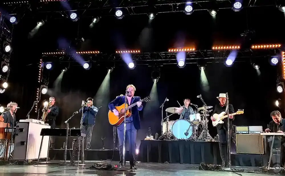 A band performs on stage with a singer playing guitar, accompanied by drums, bass, horns, and a keyboard.