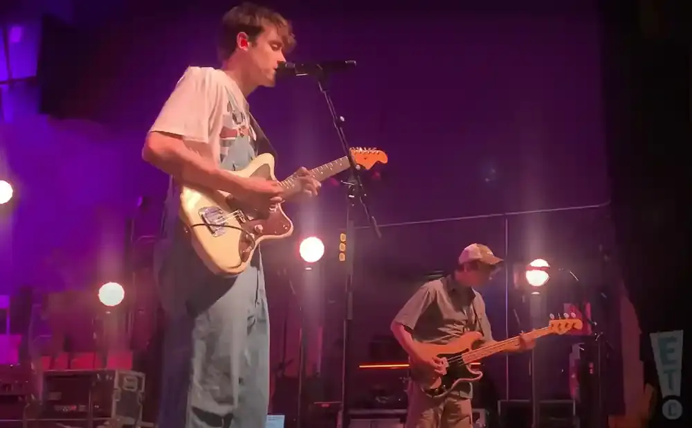 A man in overalls plays guitar on stage with a bassist in a cap.