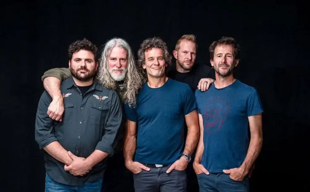 Five men are posing for a photo against a black background. The man on the far left has his arm around the man next to him.