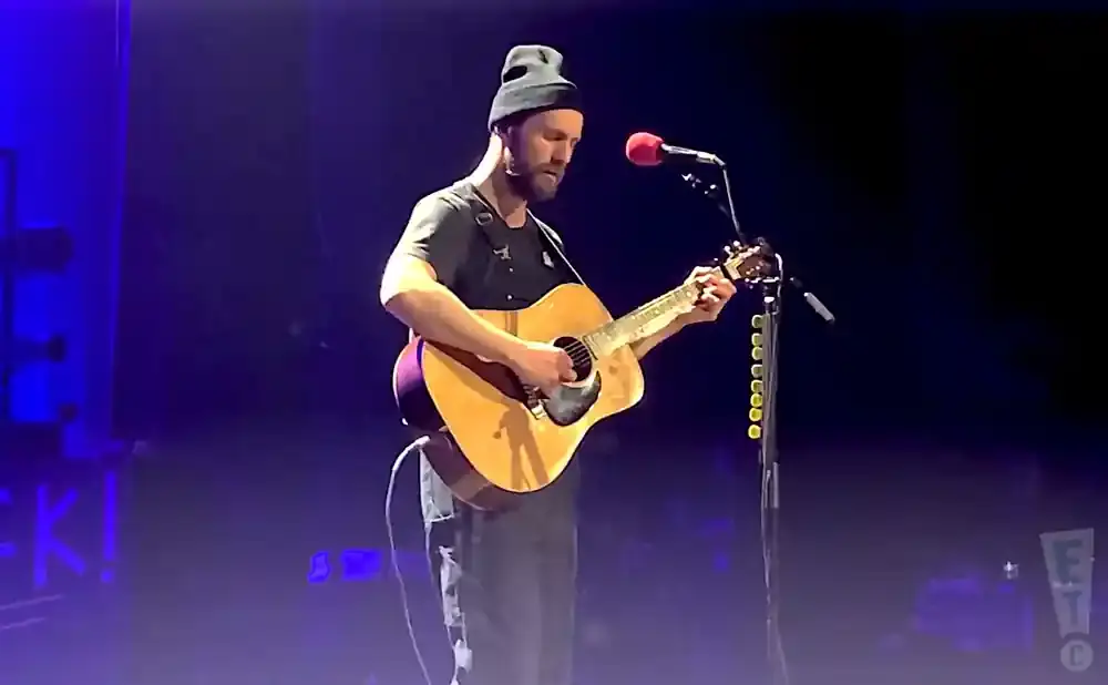 A man in a beanie plays an acoustic guitar on stage, illuminated by blue and purple lights.