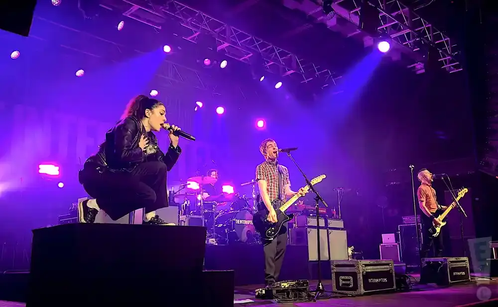 Singer crouches on a stage singing into a microphone as a band performs behind her under purple lights.