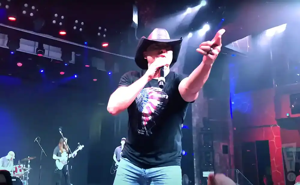 A man in a cowboy hat sings into a microphone on stage, wearing a t-shirt with an American flag design.