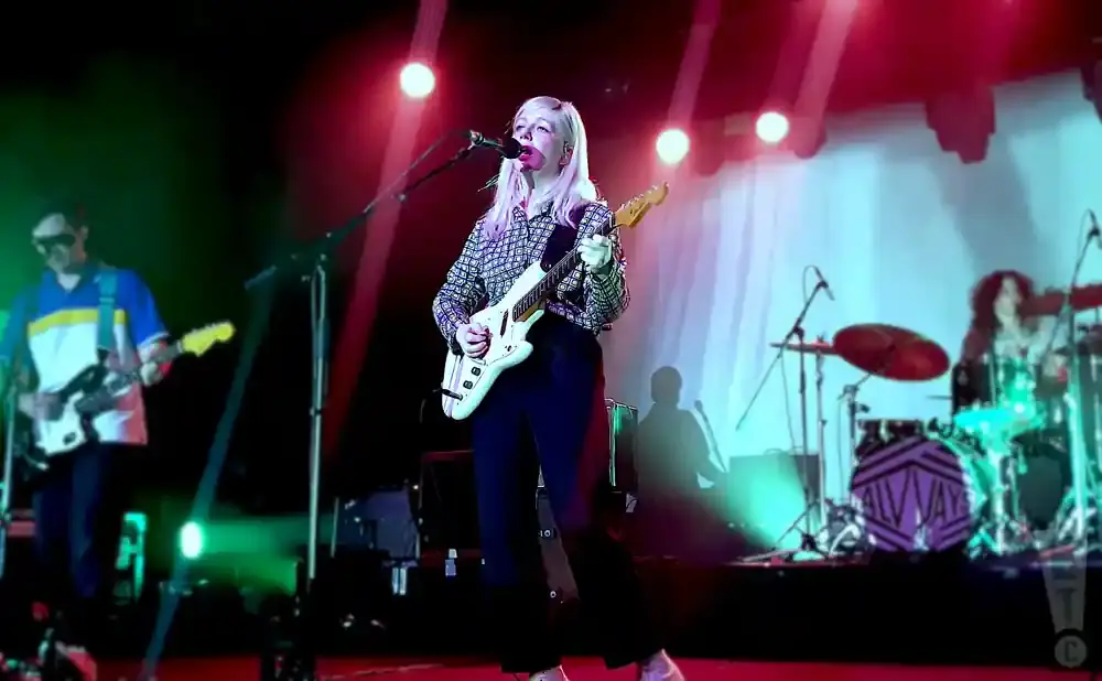 A female guitarist with blonde hair plays a white electric guitar on stage, bathed in red and green lights.