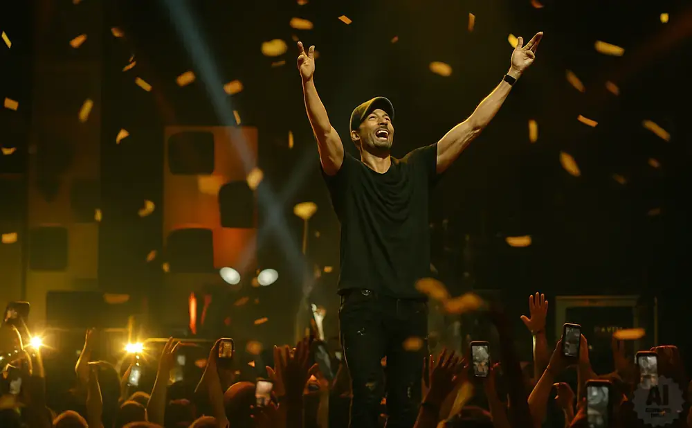 A man in a black t-shirt and cap stands on a stage with his arms raised, celebrating with confetti falling around him as a crowd cheers below.