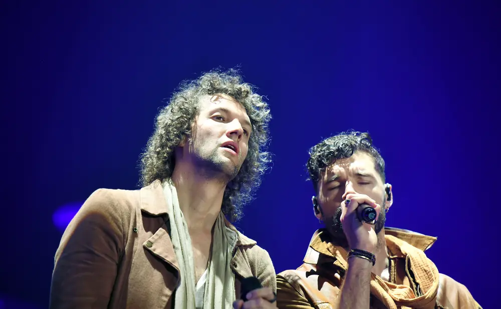 Two men singing into microphones on a stage with a blue background.