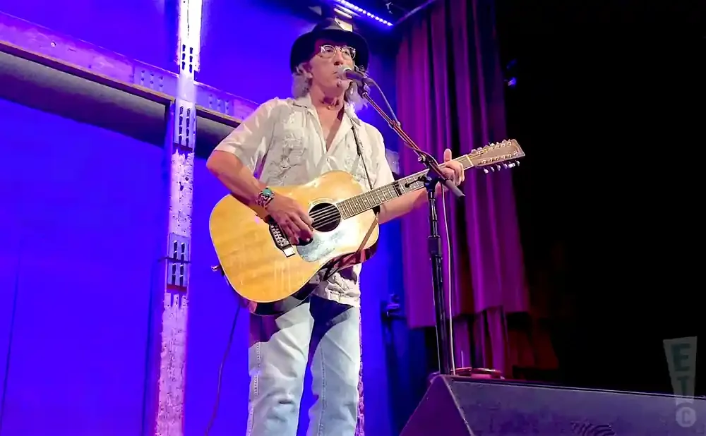 Musician plays a 12-string acoustic guitar on stage with purple curtains and blue lighting.