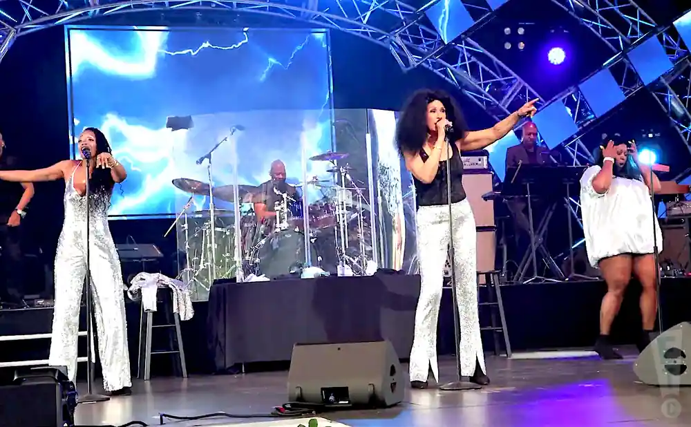 A disco band performs on stage, with a female singer in a sparkly jumpsuit in the foreground.