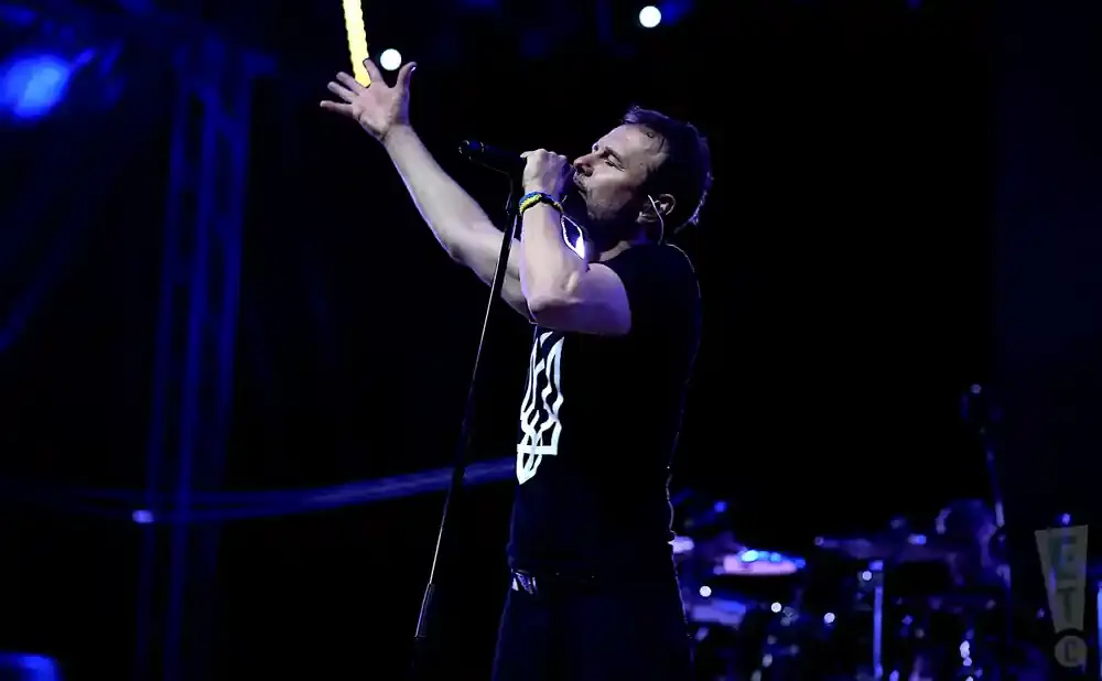 Singer on stage with microphone, wearing a black t-shirt with a white symbol and a yellow and blue wristband.