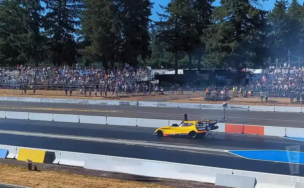 A yellow dragster speeds down a track with a crowd watching from the bleachers.