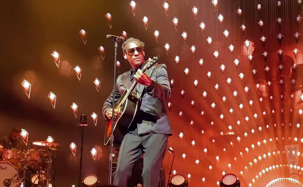 A Black man in a suit plays an acoustic guitar on stage, illuminated by diamond-shaped lights.
