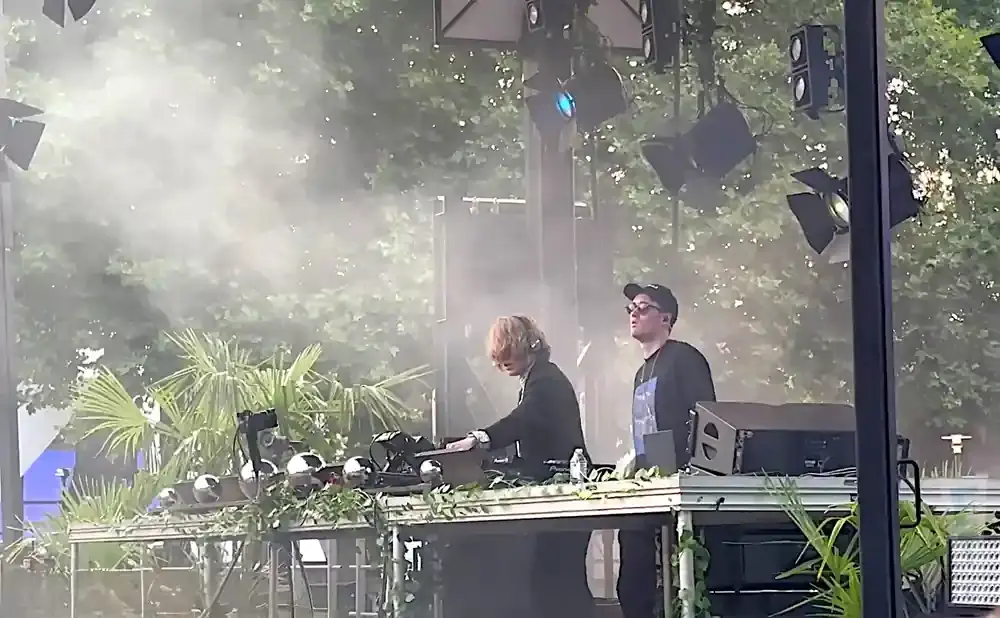 Two DJs on a stage with smoke and palm trees.
