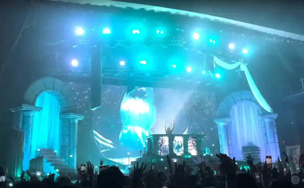 A DJ performs on a stage with a large screen displaying an icy landscape, while an audience cheers.