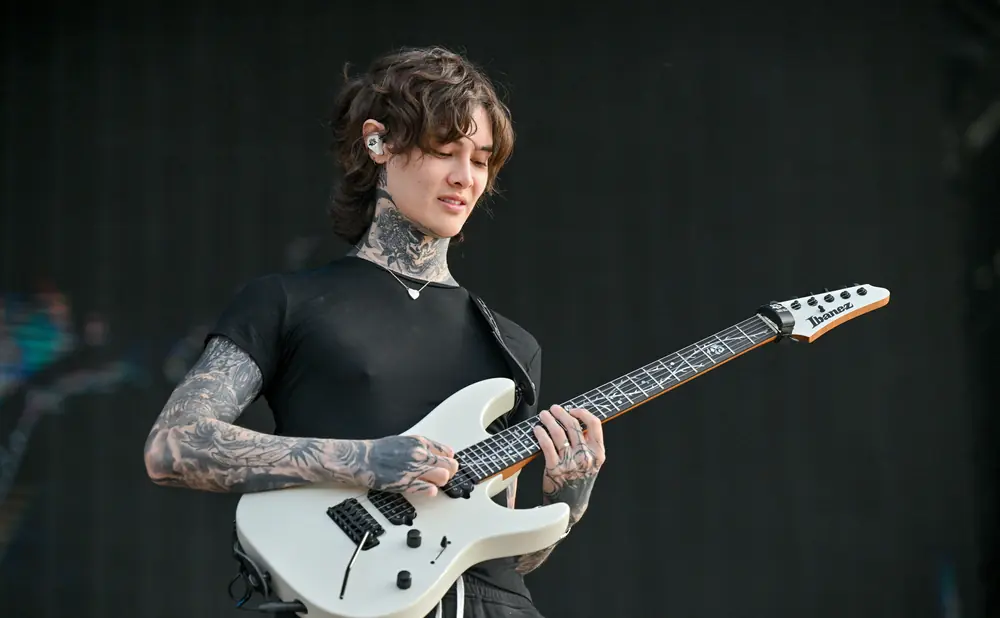 A tattooed musician plays a white Ibanez electric guitar on stage against a black background.