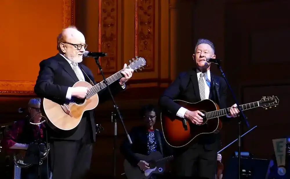 Two men in suits play guitars and sing into microphones on a stage.