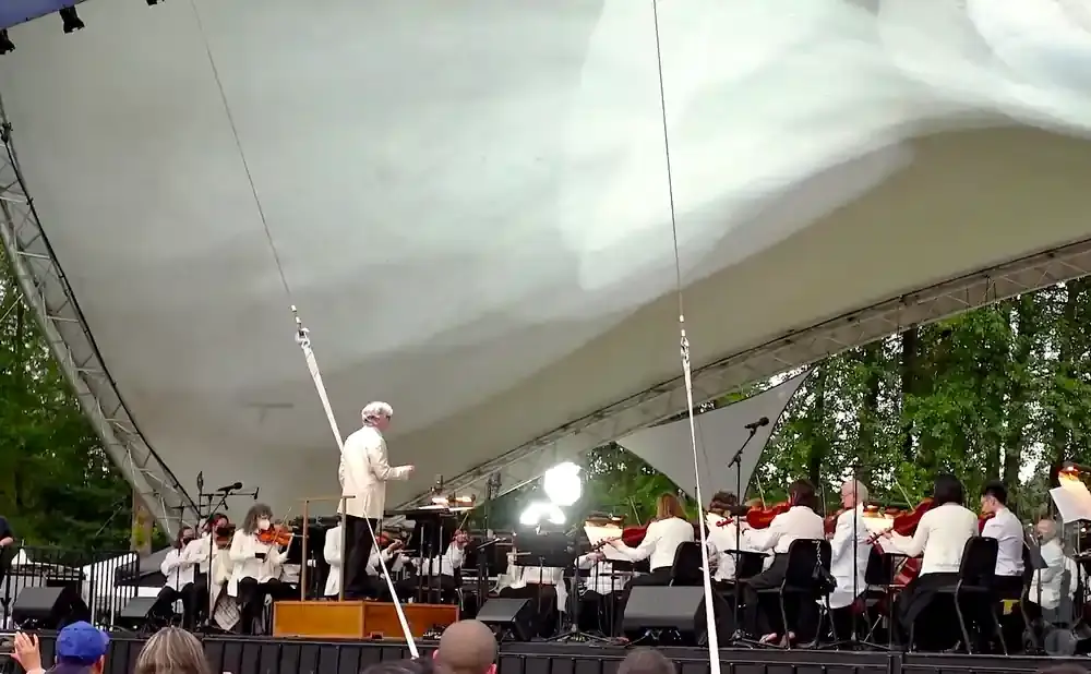 An orchestra performs under a white canopy. A conductor in a light-colored jacket leads the musicians.