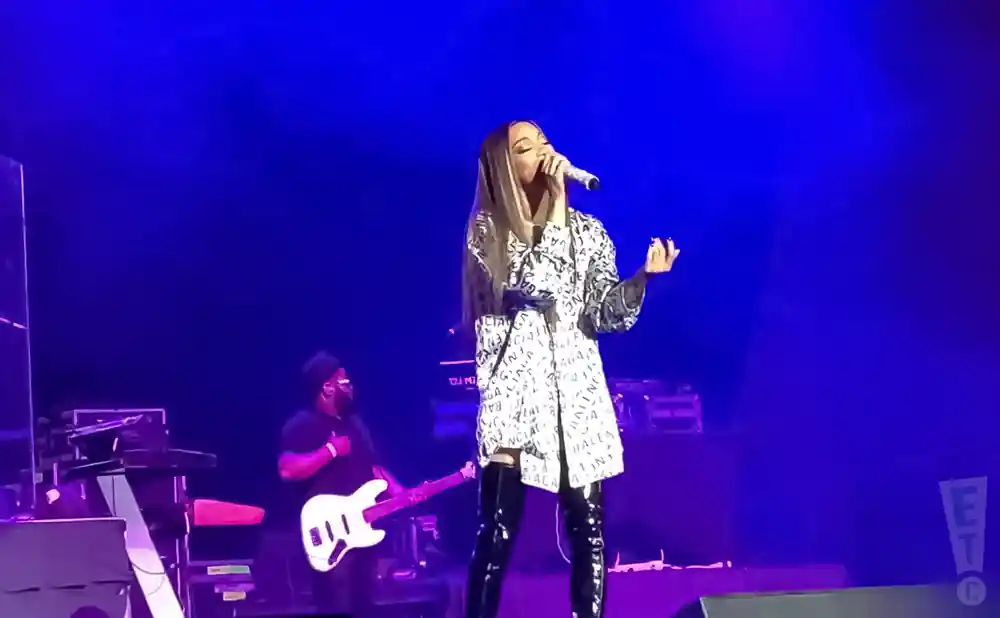 A female singer performs on a stage with blue lighting, wearing a white coat with black lettering and thigh-high boots.